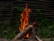 A stack of wooden logs burning with bright orange flames that rise into the air. The fire is situated in an outdoor setting, surrounded by lush green vegetation. In the background, there is a rustic wooden fence made from horizontal wooden slats, adding to the natural environment.