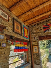 A rustic interior with stone walls and a wooden ceiling, adorned with various framed artworks and colorful flags, including Tibetan prayer flags. Natural light streams in through a large window, offering a view of an outdoor garden area where a few people can be seen walking.