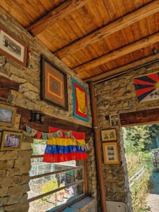 A rustic interior with stone walls and a wooden ceiling, adorned with various framed artworks and colorful flags, including Tibetan prayer flags. Natural light streams in through a large window, offering a view of an outdoor garden area where a few people can be seen walking.