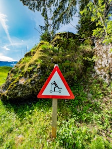 A triangular warning sign with a red border and a white center, featuring an illustration of a person falling off a scooter. The sign is placed in a lush green area with dense vegetation, shrubs, and trees. In the background, there are moss-covered rocks and a clear blue sky.