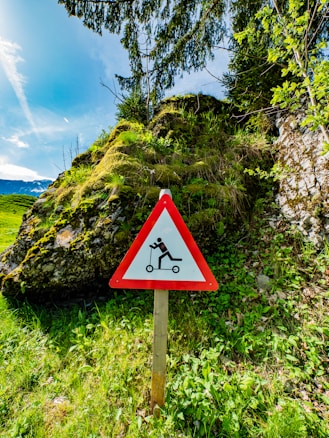 A triangular warning sign with a red border and a white center, featuring an illustration of a person falling off a scooter. The sign is placed in a lush green area with dense vegetation, shrubs, and trees. In the background, there are moss-covered rocks and a clear blue sky.