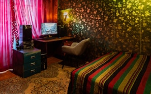 A cozy room featuring a decorated wall patterned with leaves, a comfortable chair in front of a computer desk, and vibrant red curtains covering a window. A multi-colored striped blanket is draped over a small bed, and a decorative carpet covers the floor. A painting or photo of a scenic landscape is displayed on the computer monitor.