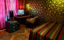 A cozy room featuring a decorated wall patterned with leaves, a comfortable chair in front of a computer desk, and vibrant red curtains covering a window. A multi-colored striped blanket is draped over a small bed, and a decorative carpet covers the floor. A painting or photo of a scenic landscape is displayed on the computer monitor.