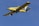 A yellow agricultural aircraft is flying against a clear blue sky. The airplane has a single propeller at the front and is equipped with spray bars under its wings.