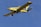 A yellow agricultural aircraft is flying against a clear blue sky. The airplane has a single propeller at the front and is equipped with spray bars under its wings.