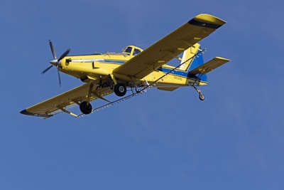 A yellow agricultural aircraft is flying against a clear blue sky. The airplane has a single propeller at the front and is equipped with spray bars under its wings.