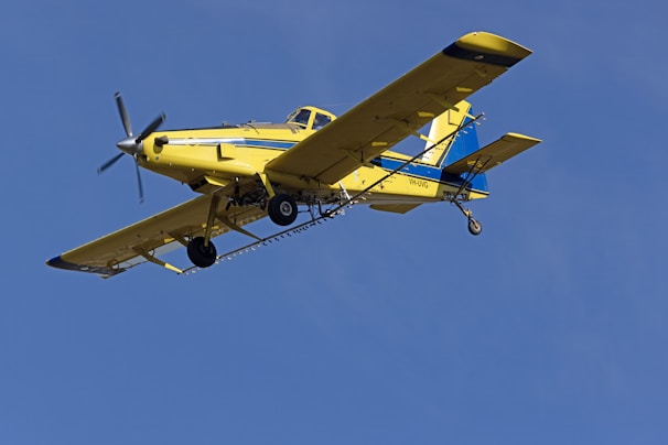 A yellow agricultural aircraft is flying against a clear blue sky. The airplane has a single propeller at the front and is equipped with spray bars under its wings.