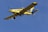 A yellow agricultural aircraft is flying against a clear blue sky. The airplane has a single propeller at the front and is equipped with spray bars under its wings.