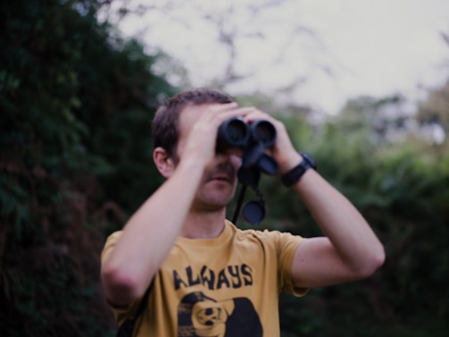A person wearing a yellow shirt is looking through binoculars in a natural, green outdoor setting.