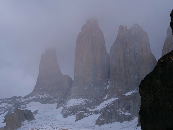 Three towering granite peaks rise dramatically against a cloudy sky, partially shrouded in mist. Snow covers the lower sections and ridges, emphasizing the rugged texture of the rock formations. The scene conveys a sense of isolation and natural grandeur.