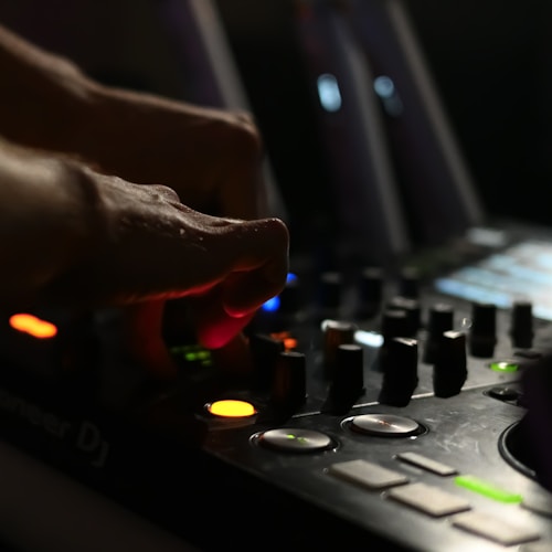 A close-up of a person's hand adjusting controls on a DJ mixer. The scene is dimly lit, highlighting the glowing buttons and knobs with different colored lights, including red, orange, and green.