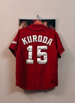 A red sports jersey with the name 'Kuroda' and the number '15' in large white letters hangs on a wall hook. The sleeves have black trim and a logo on one side. Some faces are partially visible behind the jersey, indicating that it is hanging against a photograph or poster.