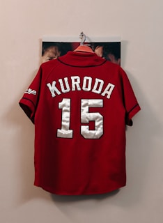A red sports jersey with the name 'Kuroda' and the number '15' in large white letters hangs on a wall hook. The sleeves have black trim and a logo on one side. Some faces are partially visible behind the jersey, indicating that it is hanging against a photograph or poster.