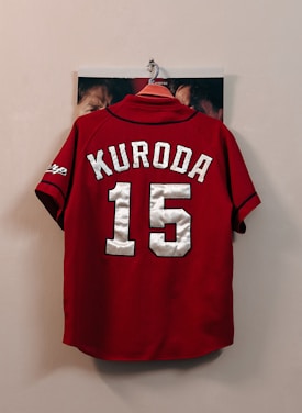 A red sports jersey with the name 'Kuroda' and the number '15' in large white letters hangs on a wall hook. The sleeves have black trim and a logo on one side. Some faces are partially visible behind the jersey, indicating that it is hanging against a photograph or poster.