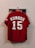 A red sports jersey with the name 'Kuroda' and the number '15' in large white letters hangs on a wall hook. The sleeves have black trim and a logo on one side. Some faces are partially visible behind the jersey, indicating that it is hanging against a photograph or poster.