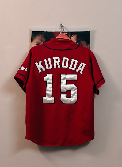 A red sports jersey with the name 'Kuroda' and the number '15' in large white letters hangs on a wall hook. The sleeves have black trim and a logo on one side. Some faces are partially visible behind the jersey, indicating that it is hanging against a photograph or poster.