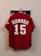 A red sports jersey with the name 'Kuroda' and the number '15' in large white letters hangs on a wall hook. The sleeves have black trim and a logo on one side. Some faces are partially visible behind the jersey, indicating that it is hanging against a photograph or poster.