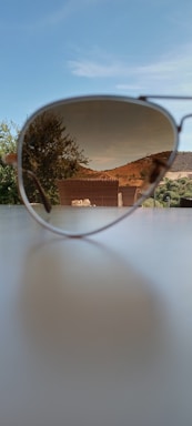 A pair of sunglasses is placed on a surface, focusing on the reflection of a scenic outdoor view in one of the lenses. The background shows a clear blue sky, a tree, and a hilly landscape with greenery.