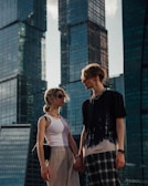 A young couple stands in front of modern high-rise glass buildings, holding hands. The woman wears sunglasses, a white tank top, beige pants, and carries a black bag. The man wears a black and white shirt with checkered pants. The setting is urban, and the light suggests either late afternoon or early evening.