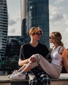 A young couple is sitting on a ledge in an urban environment with modern skyscrapers in the background. Both individuals are wearing casual clothing and sunglasses, and the sunlight creates a warm atmosphere.