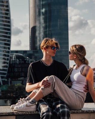 A young couple is sitting on a ledge in an urban environment with modern skyscrapers in the background. Both individuals are wearing casual clothing and sunglasses, and the sunlight creates a warm atmosphere.