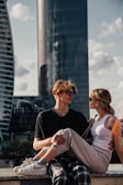 A young couple is sitting on a ledge with modern skyscrapers in the background. They are both wearing sunglasses, and the woman is sitting on the man's lap. The setting appears to be urban with a relaxed vibe, possibly during the day.