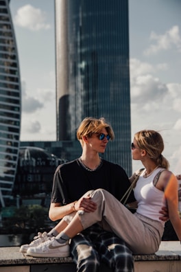 A young couple is sitting on a ledge with modern skyscrapers in the background. They are both wearing sunglasses, and the woman is sitting on the man's lap. The setting appears to be urban with a relaxed vibe, possibly during the day.