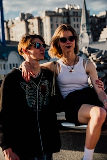 A young couple is sitting together outdoors, with buildings in the background. Both are wearing sunglasses and appear relaxed. The woman is sitting on a ledge with her arm around the man's shoulder who stands beside her. Their clothing is casual, and the setting suggests an urban environment.