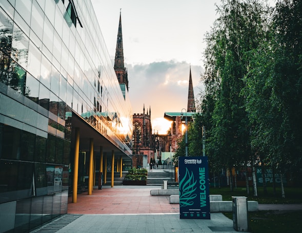 A modern architectural building with reflective glass panels is on the left, while an old cathedral with tall spires is in the center background. The foreground features a path lined with trees, and a welcome sign for Coventry University.