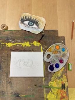 A sketchpad displays an unfinished drawing of an eye, accompanied by a detailed colored illustration of an eye on a separate piece of paper. A paint palette with various vibrant colors, a brush, a pencil, a small roll of tape, and a box of crayons are also present on the wooden table.