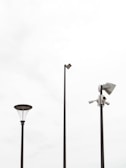 Three tall poles placed against a bright, overcast sky. One pole features a street lamp with a modern design, another has a mounted security camera setup, and the tallest pole supports a flat light fixture.