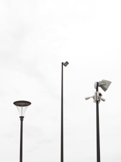 Three tall poles placed against a bright, overcast sky. One pole features a street lamp with a modern design, another has a mounted security camera setup, and the tallest pole supports a flat light fixture.