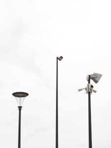 Three tall poles placed against a bright, overcast sky. One pole features a street lamp with a modern design, another has a mounted security camera setup, and the tallest pole supports a flat light fixture.