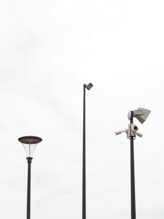 Three tall poles placed against a bright, overcast sky. One pole features a street lamp with a modern design, another has a mounted security camera setup, and the tallest pole supports a flat light fixture.