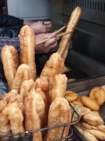 Golden-brown dough sticks and various pastries are being deep-fried in a large fryer. Several hands with chopsticks are manipulating the sticks in the oil, while a basket holds more of these fried items.
