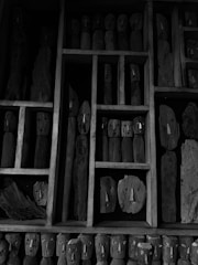 A collection of rustic, carved wooden figures organized within a grid-like wooden frame. Each small sculpture features simple carved face details, and they vary in size and shape. The figures are mostly cylindrical or oblong, giving them a somewhat abstract and primitive appearance. The artwork is monochrome, casting a somber and mysterious ambiance.