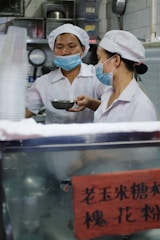 Two people wearing white uniforms and masks are working in a kitchen environment. They both wear white caps and are focused on their tasks. One of them is holding a metal bowl or container. The background includes kitchen equipment, shelves, and containers. A red sign with Chinese characters is visible.