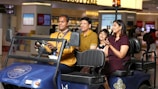 A family of three, comprising two adults and a child, is seated on the back of a blue airport buggy driven by a uniformed staff member. They appear cheerful and engaged, with the child looking particularly excited. The setting is an airport terminal with various shops and duty-free areas in the background, indicated by the visible signage and displays.