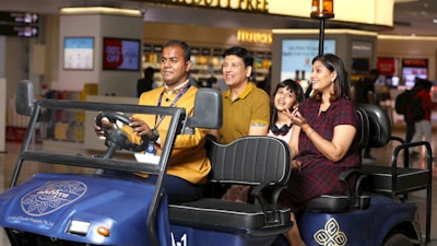A family of three, comprising two adults and a child, is seated on the back of a blue airport buggy driven by a uniformed staff member. They appear cheerful and engaged, with the child looking particularly excited. The setting is an airport terminal with various shops and duty-free areas in the background, indicated by the visible signage and displays.