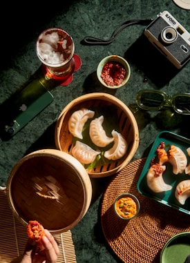 A tabletop scene featuring steamed dumplings in a bamboo steamer and on a green tray. Accompanying the dumplings are bowls of dipping sauces, a glass of beer on a red coaster, a pair of green sunglasses, a camera, a green smartphone, and a woven placemat. A hand is holding a dumpling with red chili sauce.