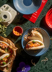 A green tabletop displaying a variety of objects including three bao buns filled with colorful vegetables on blue and red plates, a small cup of orange dipping sauce, a Polaroid instant camera, some green leaves, and a purple flower-shaped item.