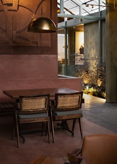 A warmly lit indoor space with a wooden table and two woven-back chairs. The room features an industrial design with metal beams and a large bronze pendant light above the table. Plants and framed artwork can be seen in the background, contributing to a cozy ambiance.