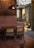 A warmly lit indoor space with a wooden table and two woven-back chairs. The room features an industrial design with metal beams and a large bronze pendant light above the table. Plants and framed artwork can be seen in the background, contributing to a cozy ambiance.