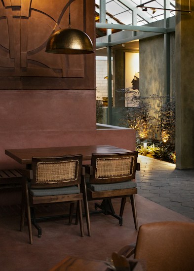 A warmly lit indoor space with a wooden table and two woven-back chairs. The room features an industrial design with metal beams and a large bronze pendant light above the table. Plants and framed artwork can be seen in the background, contributing to a cozy ambiance.