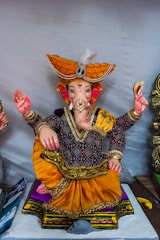 A colorful statue of a deity with an elephant head, wearing an intricately designed outfit with a vibrant orange turban and gold embellishments. The figure is sitting cross-legged, holding objects in its hands, and is adorned with detailed ornaments.