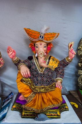 A colorful statue of a deity with an elephant head, wearing an intricately designed outfit with a vibrant orange turban and gold embellishments. The figure is sitting cross-legged, holding objects in its hands, and is adorned with detailed ornaments.
