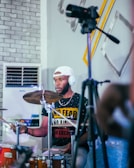 A person is playing drums while seated, wearing headphones and a cap. The background features a wall with a brick pattern and abstract art pieces, along with an air conditioner. A microphone or camera on a tripod is in the foreground.