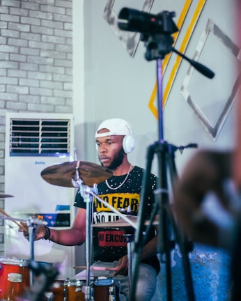 A person is playing drums while seated, wearing headphones and a cap. The background features a wall with a brick pattern and abstract art pieces, along with an air conditioner. A microphone or camera on a tripod is in the foreground.