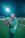 A person wearing a red bucket hat and a green shirt is taking a photograph on a sports field at night. A bright floodlight illuminates the scene. In the background, several other people are visible, wearing green uniforms, suggesting a sports event or practice.