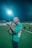 A person wearing a red bucket hat and a green shirt is taking a photograph on a sports field at night. A bright floodlight illuminates the scene. In the background, several other people are visible, wearing green uniforms, suggesting a sports event or practice.